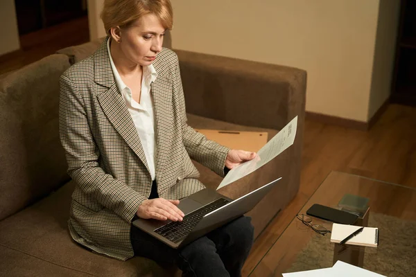 中年の働く女性はノートパソコン付きの柔らかいソファの上に座っています ビジネス女性は文書で動作します — ストック写真