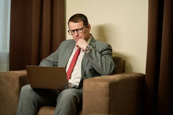 戴眼镜的严肃男性坐在沙发上 膝头靠着笔记本电脑 全神贯注地工作 — 图库照片