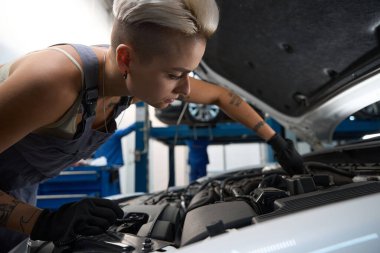 Koruyucu eldivenli güzel kadın araba tamircisi arabanın motorunu kontrol ediyor, dövmeli ve iş tulumlu kadın.