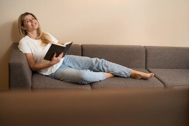 Kameraya gülümseyen ve evde kitap okurken rahat bir kanepeye uzanan mutlu kadın.