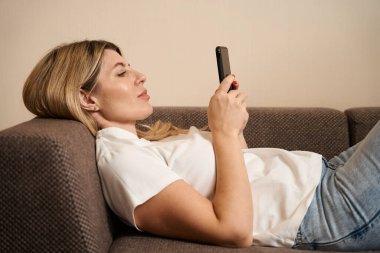 Dairelerdeki rahat kanepede dinlenirken, cep telefonuna göz atan günlük kıyafetlerle çekici bir kadın.