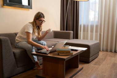 Tişört ve kot pantolon giymiş düşünceli bir kadın kanepeye oturmuş ve modern odada dizüstü bilgisayarını karıştırıyor.