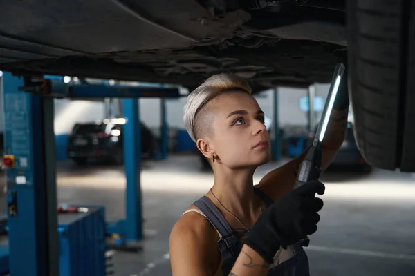 若い女性はリフトで調達車の下で修理作業を行い 彼女は特別なランプを使用しています — ストック写真