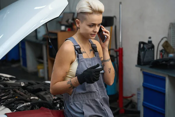 電話で話している自動車修理工場の職場の女性 職人の女性は短い散髪をしています — ストック写真
