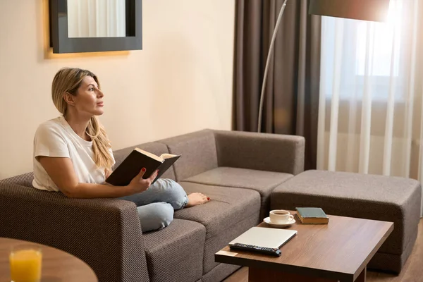 一个多愁善感的女人坐在客厅里舒服的沙发上 手里拿着书 望着别处 — 图库照片