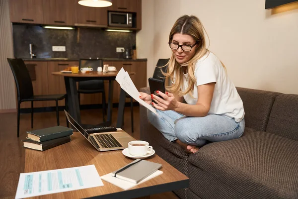 眼鏡の専門的な女性労働者は 紙を保持し キッチンでモバイルやノートパソコンを使用して課題を行う — ストック写真