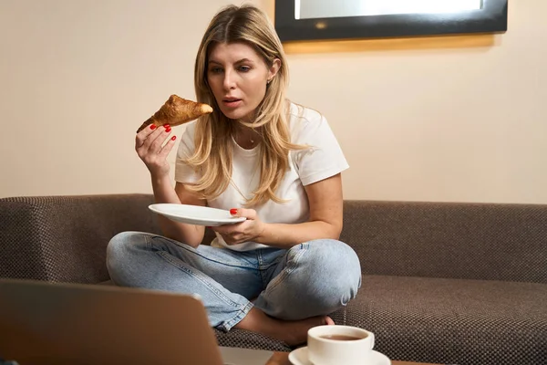 有魅力的女人穿着休闲装坐在沙发上 边喝咖啡边吃羊角面包 同时在室内使用笔记本电脑 — 图库照片