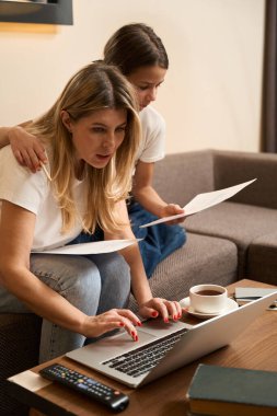 Kibar anne kanepede oturup dizüstü bilgisayarda ödevlerini yaparken kızı modern odada ona sarılıyor.