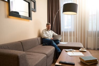 Rahat koltukta oturmuş, rahat kıyafetlerle gülümseyen adam içeride dizüstü bilgisayarla çalışıyor.