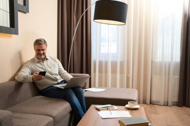 Kanepede oturan nazik bir erkek, bacaklarında defter olan ve cep telefonunu içeride gezdiren.