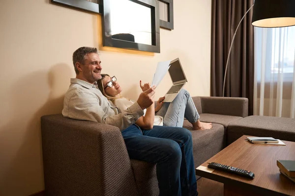 一个英俊的男人向笑着的妻子展示纸片 她戴着眼镜 膝盖上有笔记本电脑 躺在沙发上 — 图库照片