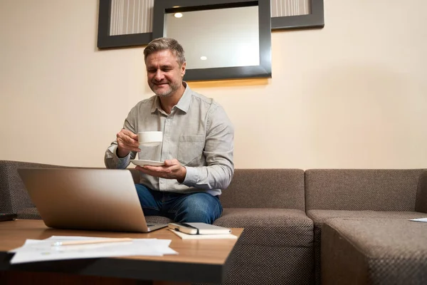 在室内使用笔记本电脑时 英俊的男性坐在舒适的沙发上 喝着一大杯咖啡来放松自己 — 图库照片