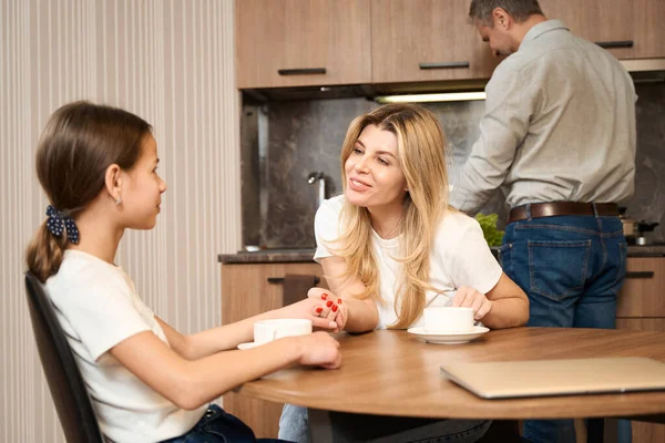 笑顔の女性は彼女の小さな娘を手に持っている 彼らはテーブルのキッチンに座ってお茶を飲んで話しています 男は背景に立って料理をしている — ストック写真