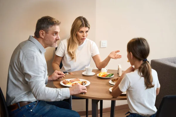 かなりの女性は Unshaven男と小さな女の子は キッチンでは テーブルに座っているとコーヒーを飲んで食べている 母と父は娘と何か不穏な話をしている — ストック写真