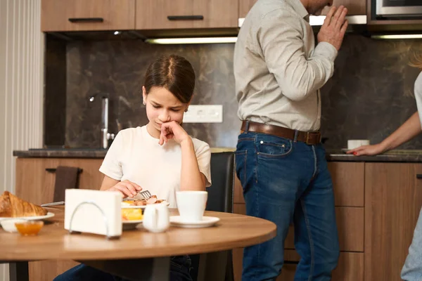 心烦意乱的小女孩正坐在厨房的桌子旁 吃喝着 她的父母正在讨论一些事 — 图库照片