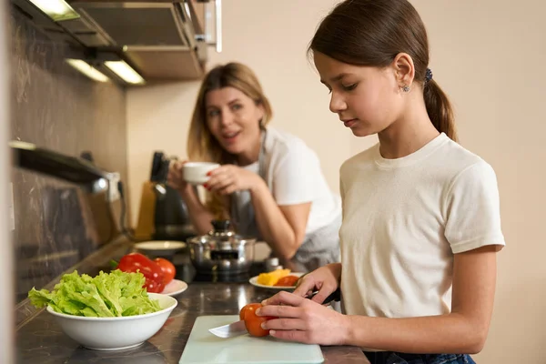 10代の娘は台所で朝食のためにトマトを切っている 母親は近くに立ってコーヒーを飲み 子供に助言をしている 台所の表面には食べ物のあるプレートがあります — ストック写真