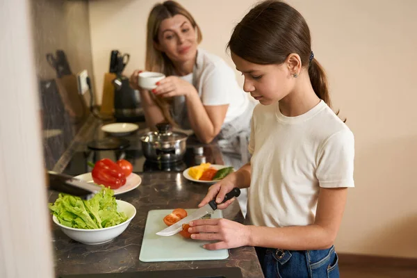 10代の女の子は台所で朝食のためにトマトを切っています 母親は近くに立ってコーヒーを飲み 子供に助言をしている 台所の表面には食べ物のあるプレートがあります — ストック写真