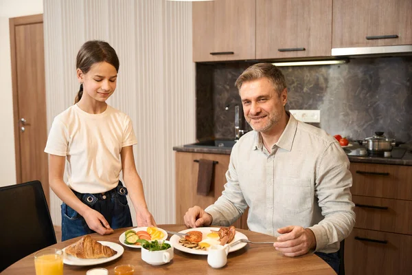笑顔の男は テーブルに座って 食べて キッチンで飲んでいます 小さな女の子が立っているとテーブルの上に食べ物とプレートを置く — ストック写真