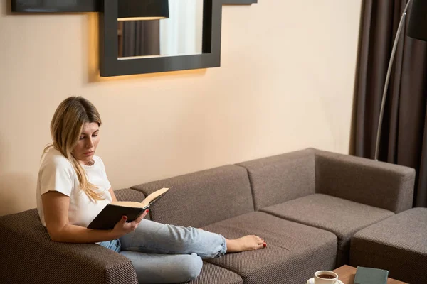 快乐的女人正一个人坐在沙发上 在旅馆房间里放松地看书 — 图库照片