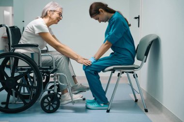 Yaşlı nazik bir hasta tekerlekli sandalyede oturuyor, bir bayan, tıp üniformalı genç bir kadını rahatlatıyor.