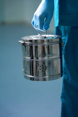 Koruyucu eldivenli ve tıbbi üniformalı bir adam hastane koridorunda parlak bir sterilizasyon kutusu taşıyor.