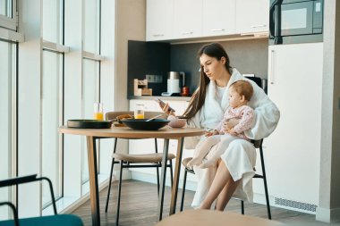 Güzel genç bir kadın mutfak masasında küçük bir çocukla oturuyor. Kadın telefondaki bir şeye bakıyor.