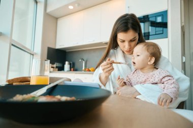 Terry bornozlu genç bir anne, bebek mamasıyla küçük bir çocuğu besler, çocuk kıyafetleri içindeki bir kızı