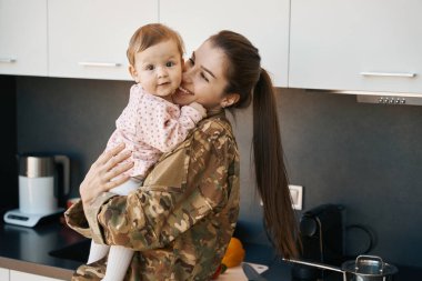 Küçük kızı kollarında tutan mutlu asker kadın, kamuflaj üniformalı anne.