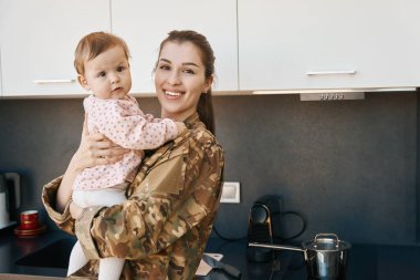 Küçük bir kızı kollarında tutan mutlu genç bir kadın, kamuflaj üniformalı bir anne.