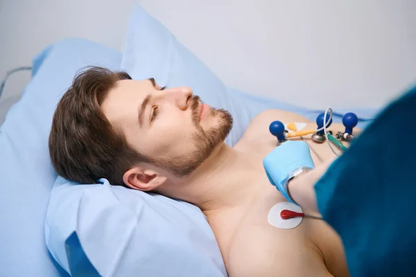 Cardiograph suction cups are attached to patient to take a cardiogram, man lies on hospital bed with a naked torso