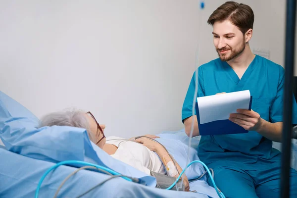Friendly Nurse Sits Patient Lying Bed Writes Her Performance — Stock Photo, Image