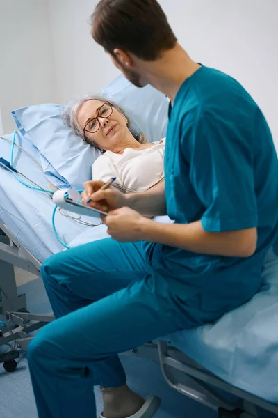 Krankenschwester Sitzt Auf Einem Bett Neben Einer Älteren Patientin Schreibt — Stockfoto