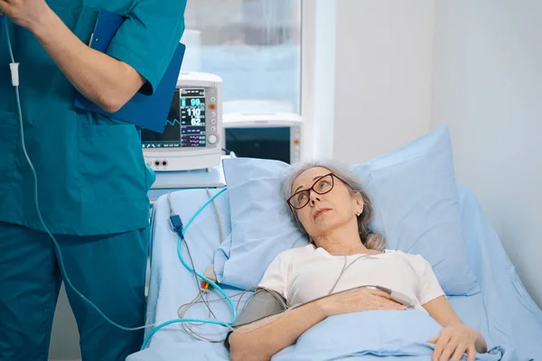 高齢者の女性は ドロッパーの下にある病院のベッドの上にあり 彼女は患者モニターに接続されており 看護師は近くにあります — ストック写真
