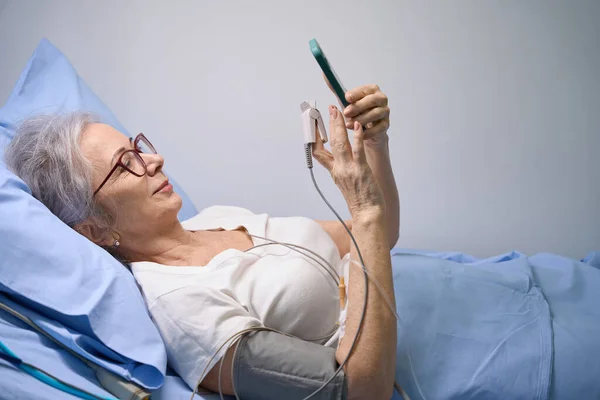 患者の回復は病院の部屋から携帯電話でチャットしている彼女は患者モニターに接続されている — ストック写真