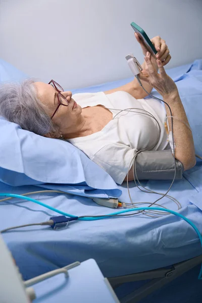 高齢の女性は病院の部屋から携帯電話でチャットしている 彼女は患者のモニターに接続されています — ストック写真