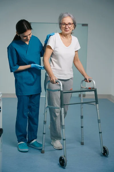 一位穿着医疗制服的年轻女性在医院走廊上陪着一位散步的老人帮助她 — 图库照片