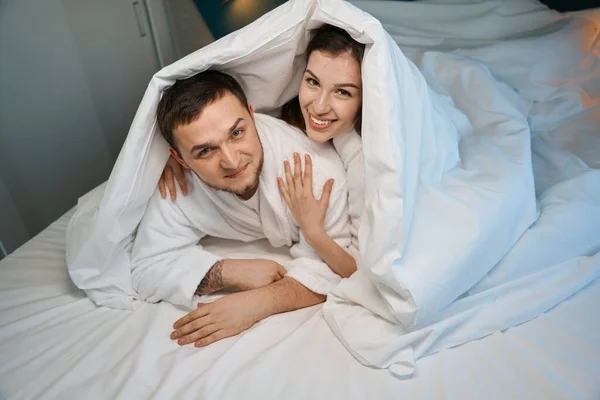 幸せな若いカップルは大きなベッドの上で抱き合って 男たちは毛布から小屋を建てた — ストック写真
