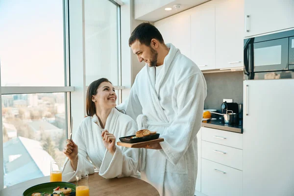 親切な男は居心地の良い雰囲気の中で朝食で彼の魅力的な妻を扱い カップルは大きなビューウィンドウに位置しています — ストック写真