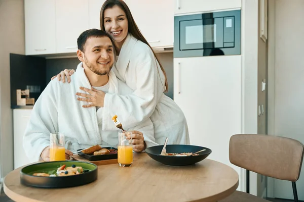 一对穿着浴袍的年轻夫妇在舒适 简约的厨房里吃早餐 一个女人温柔地拥抱一个男人 — 图库照片