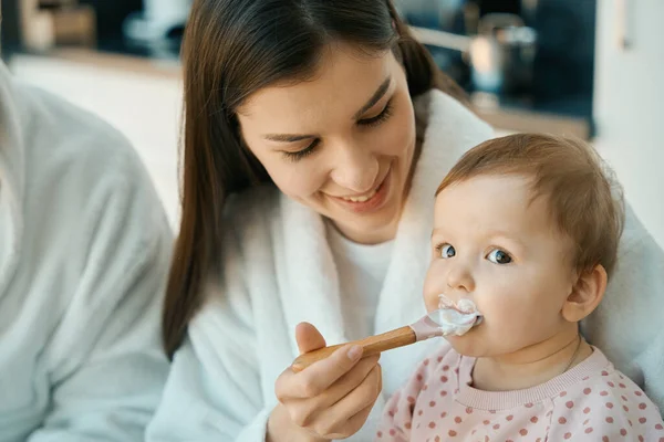 幸せな女性は小さなスプーンで赤ちゃんを食べます 女の子はふっくら頬を持っています — ストック写真