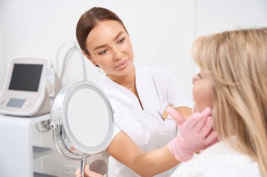 Doktor kozmetik uzmanı bir estetik tıp kliniğinin müşterisine danışıyor, bir kadın aynaya bakıyor.
