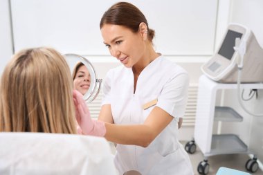 Estetik tıp kliniğindeki kozmetik uzmanı bornozlu bir hastaya, tıbbi üniformalı bir çalışana danışıyor.