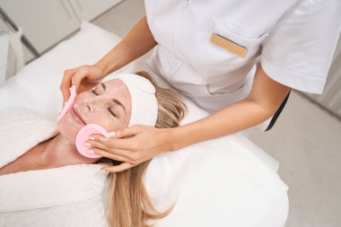 Kozmetik uzmanı, özel süngerleri olan bir kadın için canlandırıcı bir maske çıkarıyor. Hasta bu işlemden zevk alıyor.