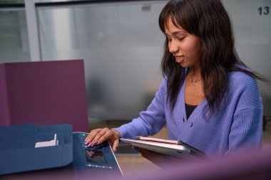 Elinde dizüstü bilgisayarla gülümseyen kadın modern odadaki çok fonksiyonlu yazıcıyı açıyor.