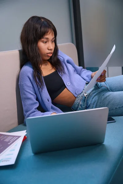 現代の部屋で居心地の良いソファに座っているときに文書を見てノートパソコンを持つ若い女性起業家 — ストック写真