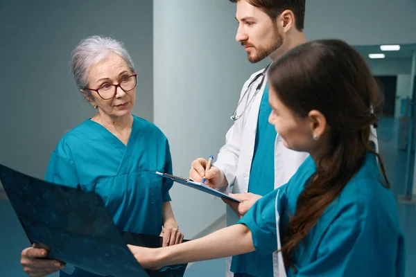 医師のグループは 診断部の廊下に立っている高齢者の女性は彼女の手にMriスキャンを持っています — ストック写真