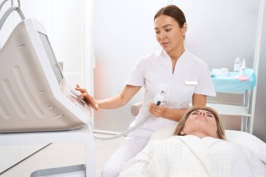 Nitelikli kadın uzman aletleri kaldırma işlemine göre ayarlar. Bir hasta yakınlardaki kanepede yatar.