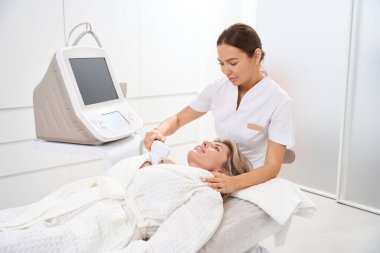 Genç bir güzellik uzmanı, bir hastanın başında oturuyor. Bir uzman, dekolte bölgesini temizliyor.