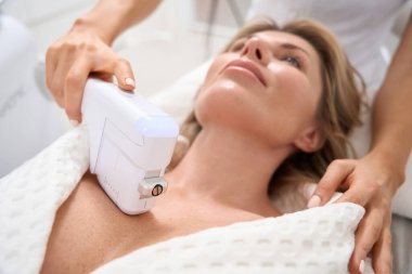 Hasta, dekolte bölgesinde cerrahi olmayan bir cilt sıkma işlemi geçiriyor. Güzellik uzmanı modern bir cihaz kullanıyor.