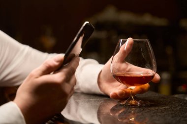Bar tezgahında tek başına otururken elinde akıllı telefon ve bir bardak viski tutan adama yaklaş.